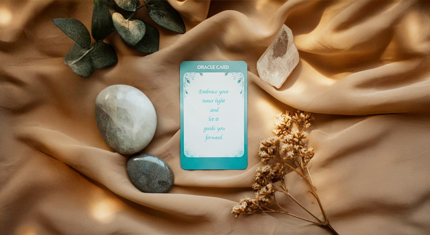 Oracle card with the message 'Embrace your inner light and let it guide you forward,' surrounded by stones, crystals, and foliage on a soft fabric background.