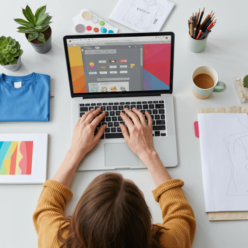 Overhead view of a creative workspace with a laptop displaying design software, surrounded by a T-shirt sketch, colorful prints, art supplies, and a coffee cup.