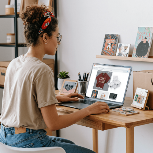 Person with glasses and headband working on a laptop at a wooden desk, displaying a t-shirt design, with art prints, a plant, and stationery in the background